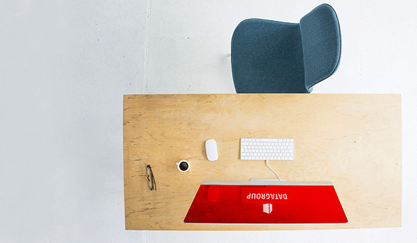 Een minimalistische werkplek met een houten bureau, een blauwe stoel en een wit toetsenbord. Op het scherm is een rode achtergrondafbeelding te zien met het DATAGROUP-logo.
