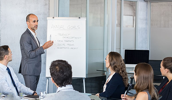 A business meeting in a modern office, during which a man presents the annual goals on a whiteboard to a group of colleagues seated around a table.